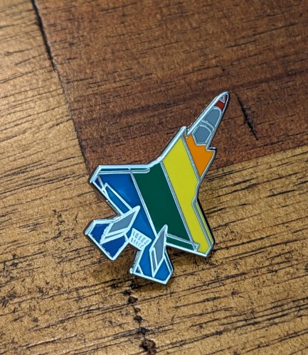 Small F-35 fighter jet enamel pin with rainbow pride flag color stripes across the jet.