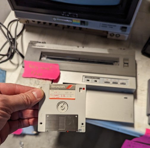 A photograph of a left hand holding a Sharp Pocket Disk, a 2½-inch variant of a hard-cased floppy disk.  It looks very similar to a 3½-inch floppy, but the longer you look at it the weirder the discrepancies get.