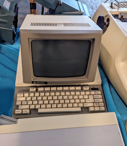A photograph of a small, old computer terminal, beige case and black screen, resting on a blue cloth.  At the bottom left of the terminal is a badge that reads, "TeleVideo Personal Terminal".