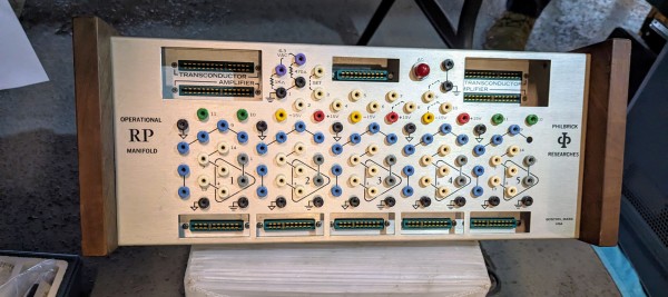 A photograph of some sort of electronics interface equipment.  A wide brushed steel face plate is set between two dark brown wooden panels.  Set in the steel face place are dozens of round terminal jacks in every color of the rainbow, with lines connecting or grouping some jacks and electrical circuit diagram signals next to others.  Above and below the field of jacks are wide green female connectors, some of which are labeled "TRANSCONDUCTOR" or "AMPLIFIER."  To the left of the jacks are the words "OPERATIONAL RP MANIFOLD."  To the right are "PHILBRICK RESEARCHES," with the words separated by a logo.  At the bottom right are the words, "BOSTON, MASS USA."  