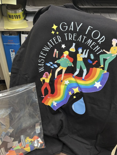 A black T-shirt with a picture of four people dancing along a rainbow-colored path. The text says Gay For Wastewater Treatment. There are also smaller images of test tubes, fish, a water droplet, etc.