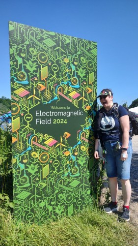 An excited pasty dork stands next to a large sign reading "Welcome to Electromagnetic Field 2024"