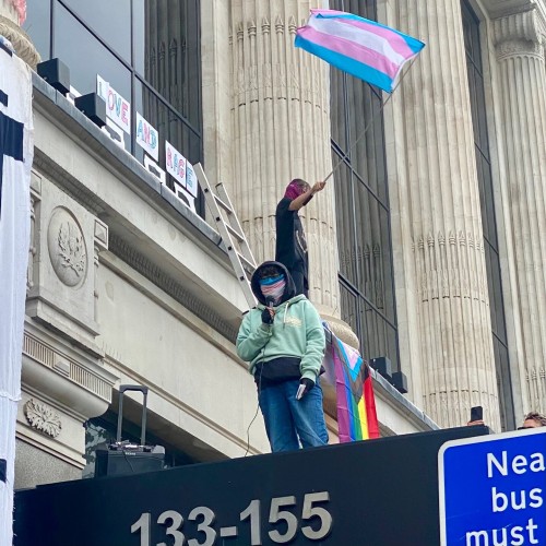 A short protester in glasses, a trans flag bandana, and a pale green sweatshirt over a grey hoodie speaks into microphone up on the awning. Up on the ledge in the background another protester waves a trans flag”