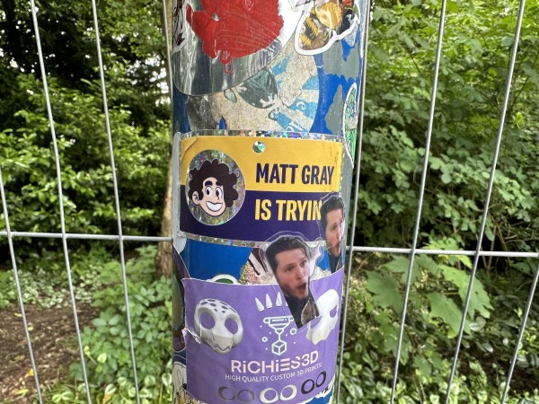 A lamp post covered in various stickers, the centre of which is a glittery sticker for the “Matt Gray is Trying” YouTube series. 