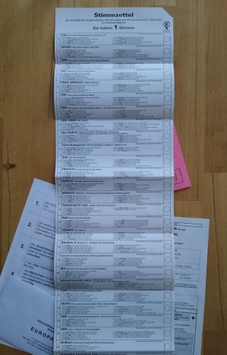 Ein auf einem Holztisch ausgebreiteter Wahlschein für die Wahl zum Europäischen Parlaments 2024, dahinter Briefumschläge und die übrigen zugehörigen Unterlagen.