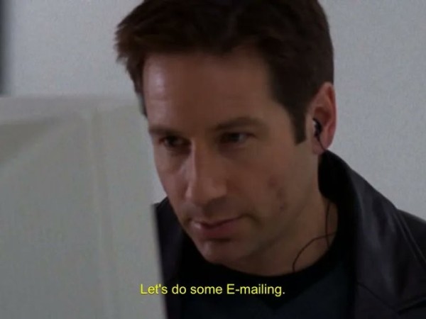 Still image. David Duchovny, a person wearing a dark blue v-neck over a black t-shirt, and black leather jacket, earpiece in left ear, looking at a CRT monitor. 

Bottom text in subtitle yellow reads:
"Let's do some E-mailing."