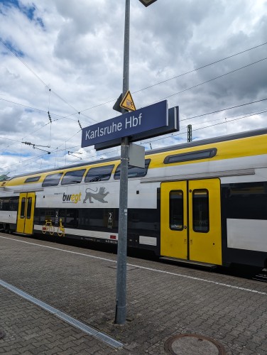A sign on a train station platform saying "Karlsruhe Hbf" with white and yellow train behind it.