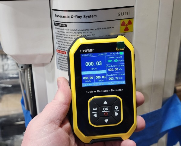 A Geiger counter reading 0.03 uSv/s held in front of a piece of medical equipment, which is labeled with the radiation symbol and "Panoramic X-ray System" 