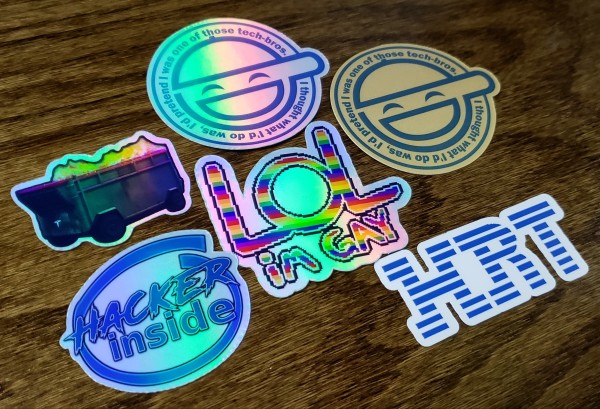 A pile of 6 stickers on a wood surface. 2 of them are of the Laughing Man from Ghost in the Shell but they say "I thought what I'd do was, I'd pretend I was one of those tech-bros.". One Laughing Man is on gold sticker paper and the other is on holographic. There is also a sticker that reads "LOL im GAY" in rainbow pixel art text. One is reminiscent of the old IBM logo but it says HRT instead. Another sticker is reminiscent of the Intel Inside logo, but it says Hacker Inside. The final sticker is a dumpster that is on fire with 4 wheels and the Tesla hood ornament.