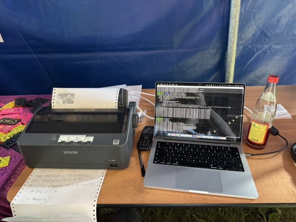 Dot matrix printer sat next to a MacBook with debug output on it.  All on a trestle table in a blue marquee