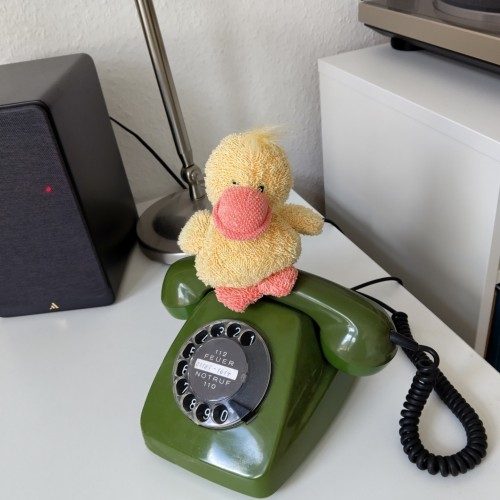 Grünes Wählscheibentelefon. Obenauf sitzt eine gelbe Kuscheltierente.