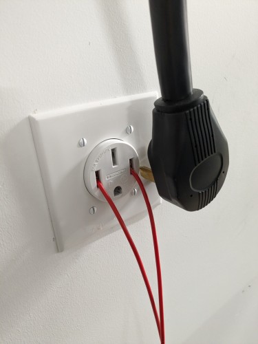 A NEMA 14-50 high power receptacle with its matching plug removed and off to the side. Two red wires are shoved on ceremoniously into the receiving pins of the receptacle and lead off frame