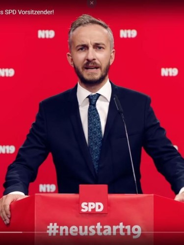 Auf dem Foto steht Jan Böhmermann im Anzug an einem roten Rednerpult, vor roter Wand. Auf der Wand ist mehrfach der Schriftzug N19 verteilt, auf dem Rednerpult steht SPD #neustart.