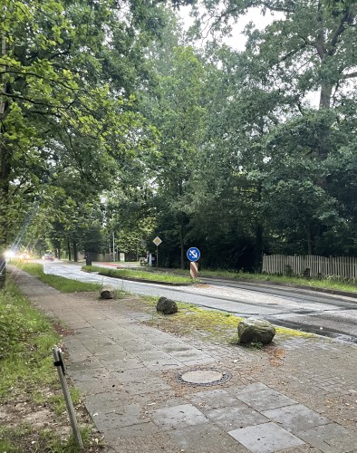 A pedestrian island on an exurban road. A sidewalk runs along the left side of the road, it is flanked by trees. Part of a residential fence is visible on the opposite side of the road.