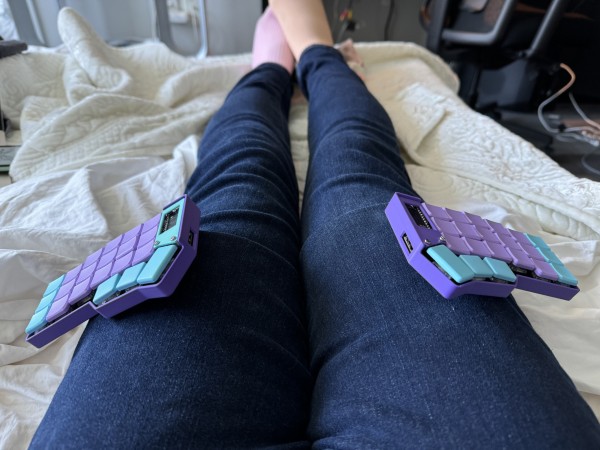 A photo of someone’s legs wearing blue jeans lying in bed. There are two small split keyboard halves, each attached to a thigh. The keyboards are small, light purple with light blue accents, and have low profile switches.