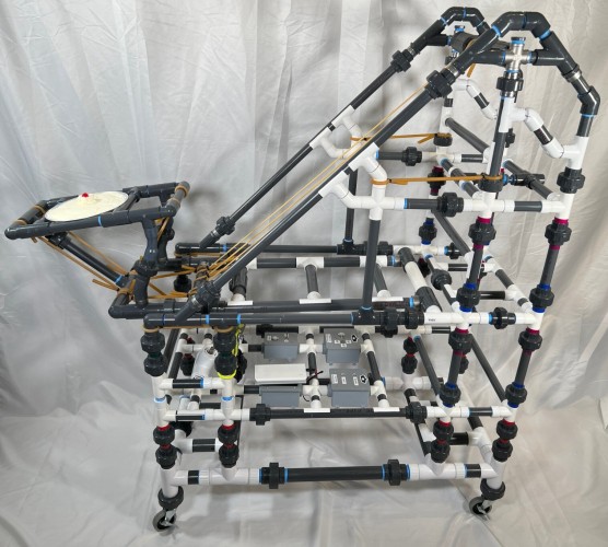 A pie launcher. It's made of a bunch of PVC pipes, building a roughly rectangular-triangular structure. There's a pie surface on the left, then some rubber tubing in the middle. The left part levers up like a catapult to launch the pie to the right.
At the bottom of the structure (which has wheels), there's several grey boxes with buttons and switches on them, for enabling the system, using a laser (for targeting?) and to enable IoT functionality. 