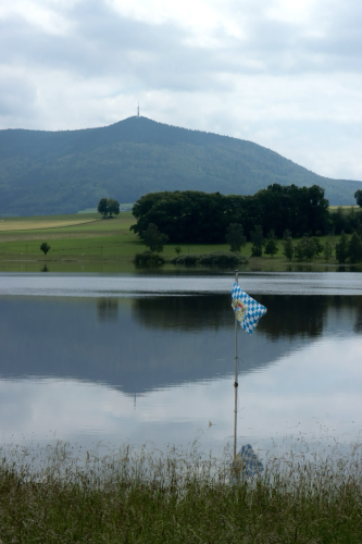 Ein See in dem eine Flagge des Freistaat Bayern weht. Im Hintergrund eine Bergkette