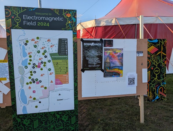 A map of the EMF campsite with all the villages and important points on it. Next to it there's a bulletin board with two posters. One of them advertises "Håck ma's" with a colourful painting of a castle on it.