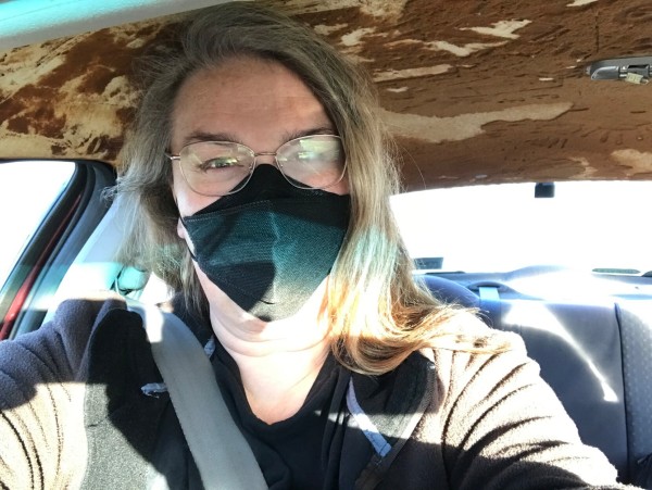 A photo of me, a youngish 53-year-old feral woman in a car with a mask on because co-worker carpooling doesn't wear one and gets frickin covid ALL THE TIME but I do ok and have avoided it so far. I look marginally better than the roof lining which has fallen off and is scattering foam and shit all over the place.