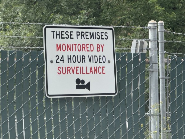 Sign on a chain-link fence reads "THESE PREMISES MONITORED BY 24 HOUR VIDEO SURVEILLANCE" with an icon of a video camera. The icon is bizarre — a long rectangle, a triangle on the end presumably a lens, and two circles on top as if it’s a reel-to-reel camera.