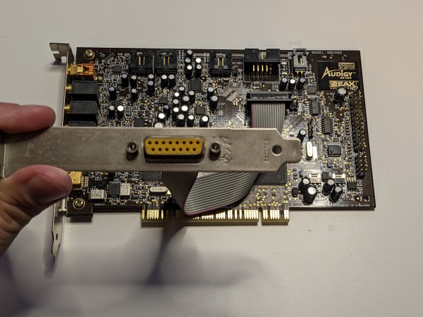 A photo of an old PC soundcard. It has a number of audio sockets on its backplate, but also has a second backplate connected by a flat ribbon cable - this backplate is being held up to the camera to show off a 15-pin socket, which is the old PC gameport socket.