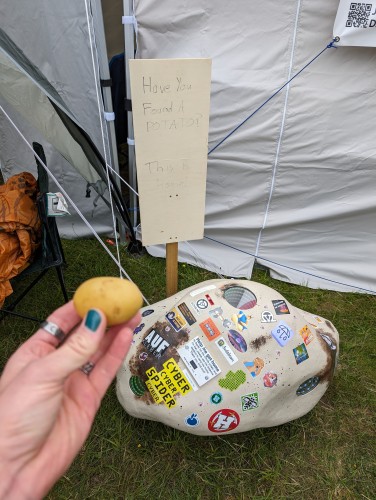 A big, potato-like thing, covered with stickers is lying on the grass in front of a sign. It's barely readable and says "Have you found a potato? This is Home!" A small potato is held in front of it.