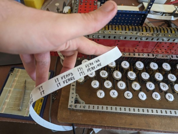 A length of ticker tape held above the keyboard of a machine built from Meccano.
A duplicated row of text is printed on the tape, which reads "IT VERKS   ITS VERKINK"