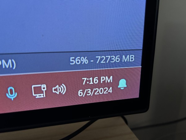 A close-up photo of a computer monitor screen showing the bottom right corner of the taskbar. The taskbar is colored red and displays icons for microphone, network, sound, and notifications, along with the date and time "7:16 PM, 6/3/2024." Above the taskbar, there is a blue section displaying memory usage statistics, indicating "56% - 72736 MB."